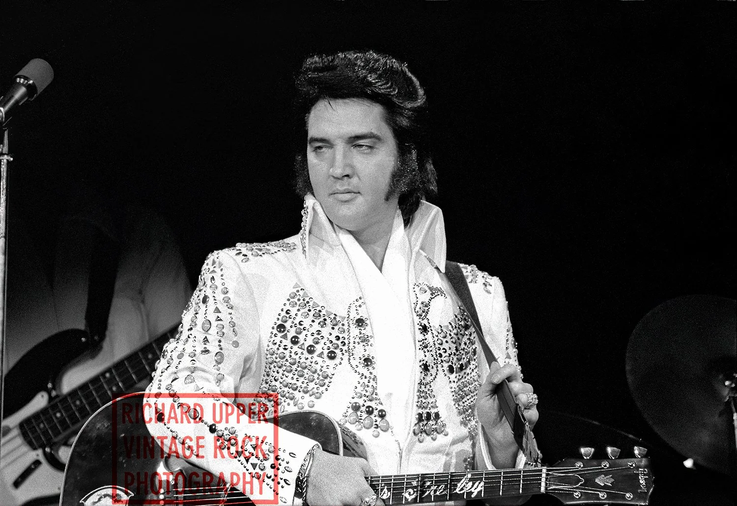 Black and white photo of Elvis Presley performing with a guitar, wearing a rhinestone-studded jumpsuit, with a microphone and drums in the background.