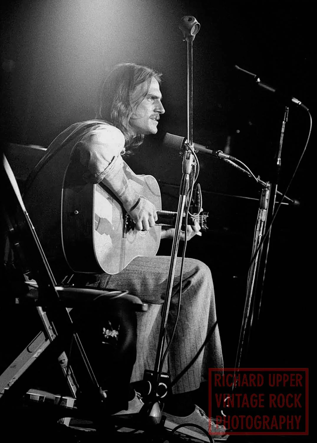 Black and white photo of a man with long hair playing an acoustic guitar on stage, with multiple microphones around him, and a spotlight shining on him, taken by Richard Upper Vintage Rock Photography.