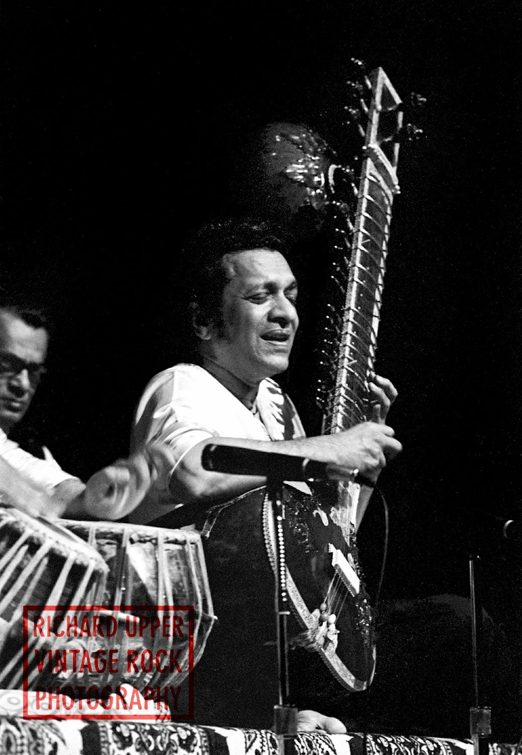 A black-and-white photo of a man playing a sitar on stage, with another musician to his left and a patterned cloth on the table in front. The watermark on the bottom left reads "Richard Upper Vintage Rock Photography."