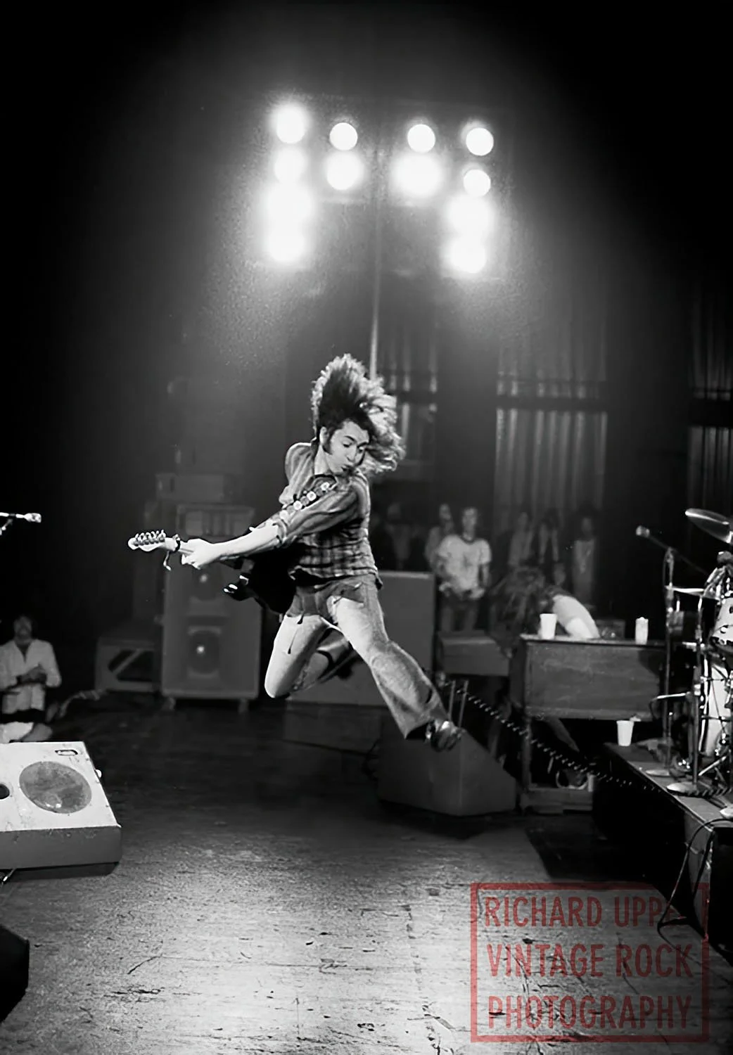 A female guitarist performing on stage, mid-air jump with a guitar, vintage-style setting, black and white photograph.
