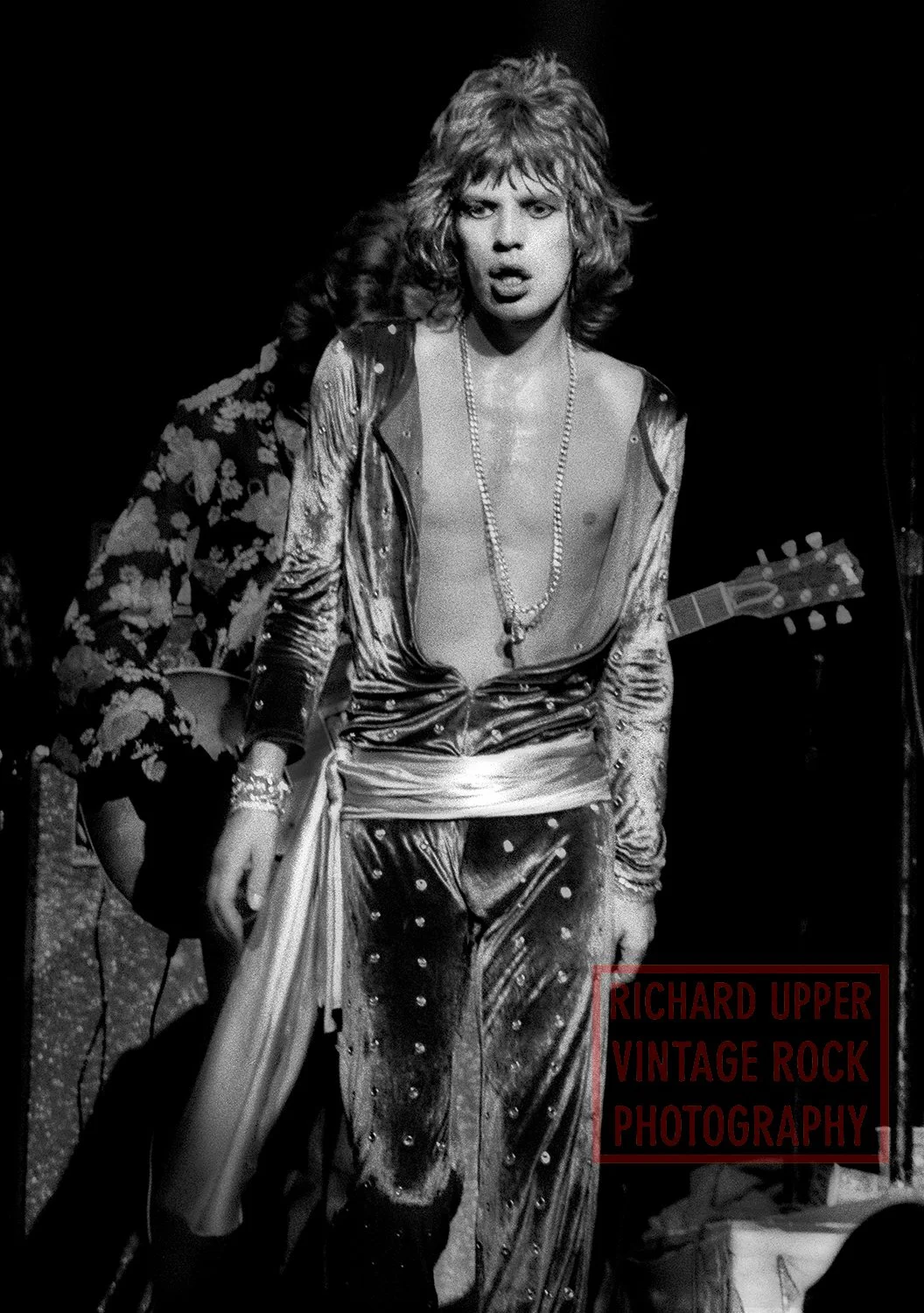 Black and white photo of a shirtless man with curly hair, wearing shiny pants and a long necklace, standing on stage with a guitar in the background, looking directly at the camera.