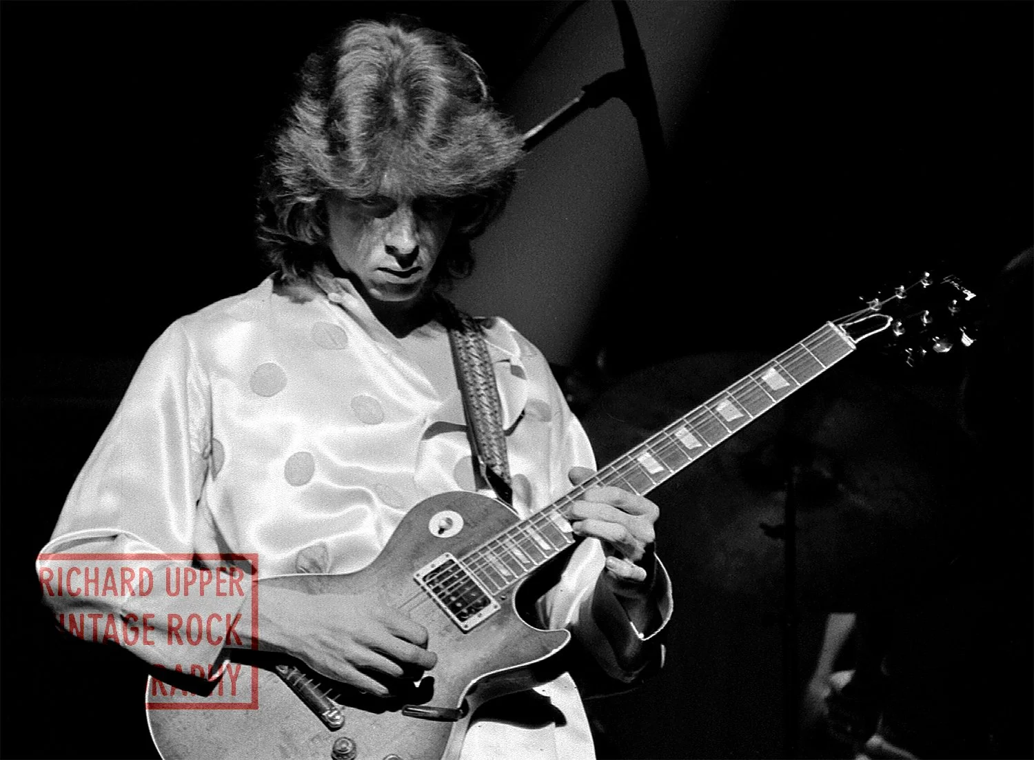 Black and white photo of a guitarist playing an electric guitar, wearing a satin shirt with polka dots, in a dark setting.