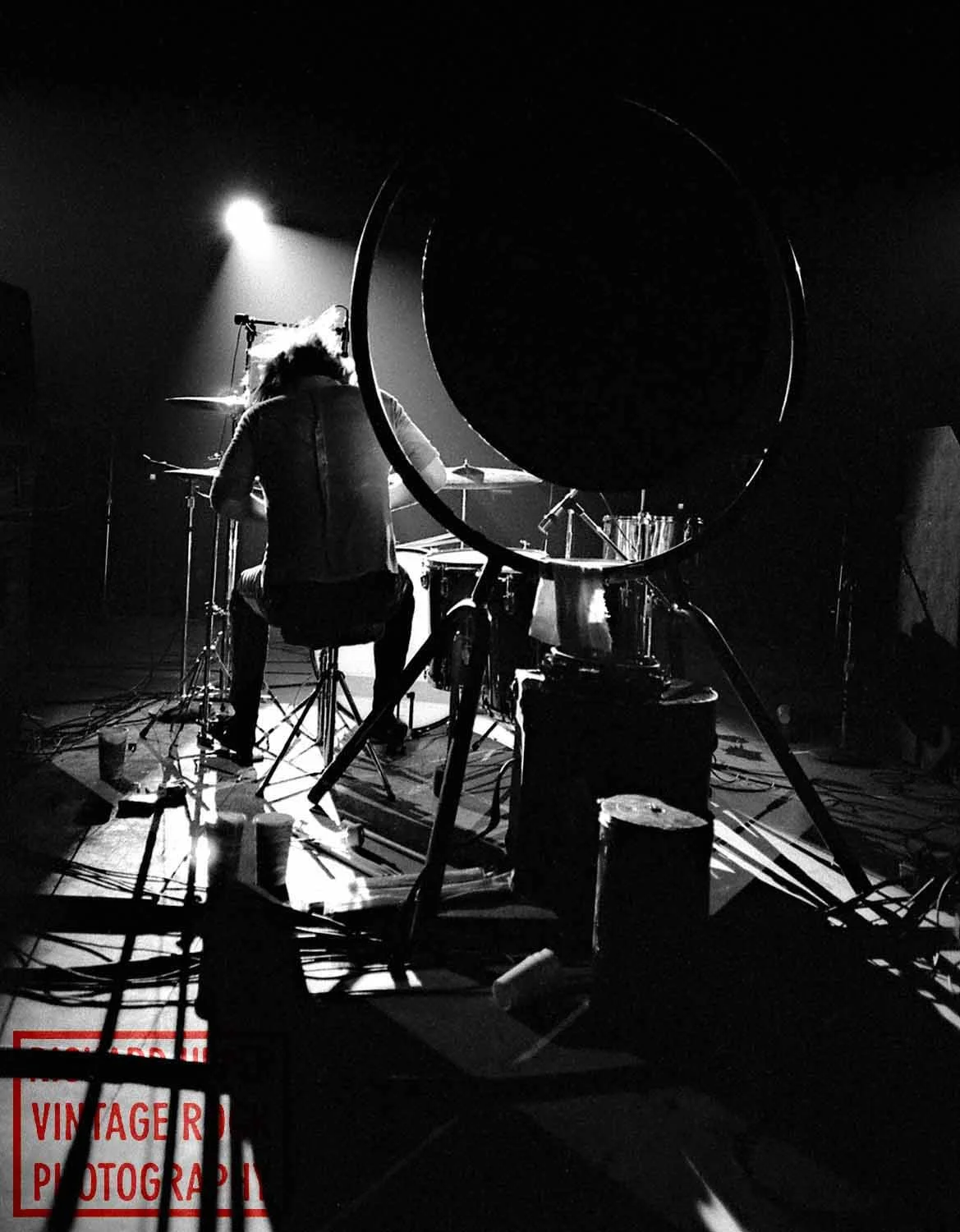Black and white photo of a musician playing drums on stage, with a bright spotlight shining behind them. Various equipment and cables are scattered on the floor.