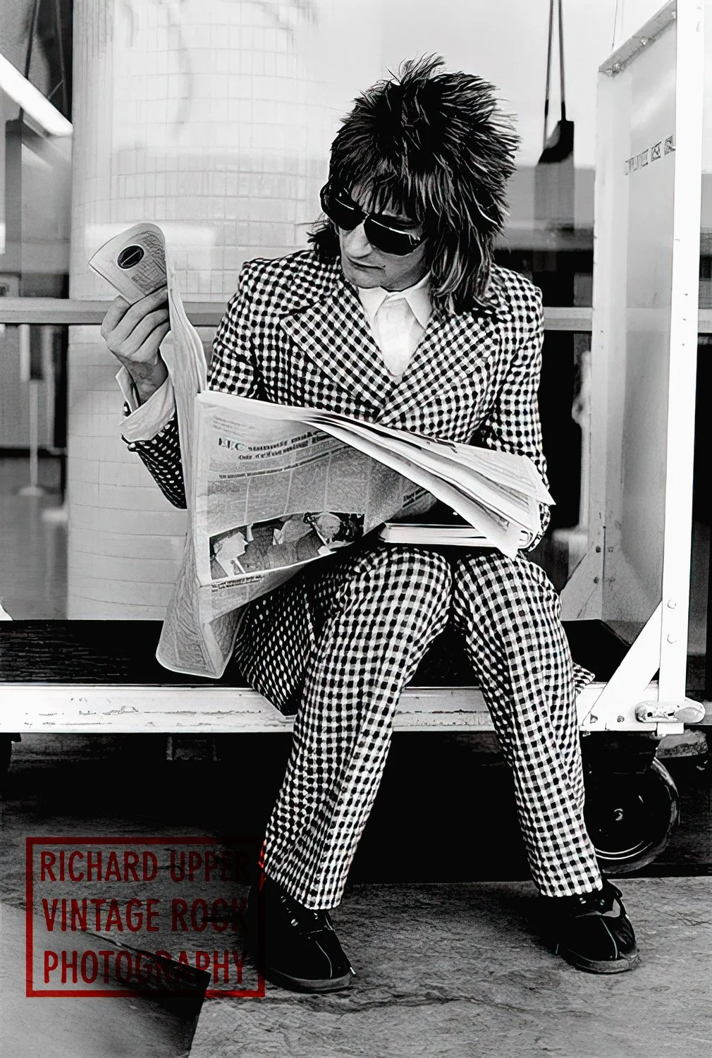 A person with spiky hair, wearing sunglasses and a checkered suit, sits on a bench reading a newspaper in an urban setting.