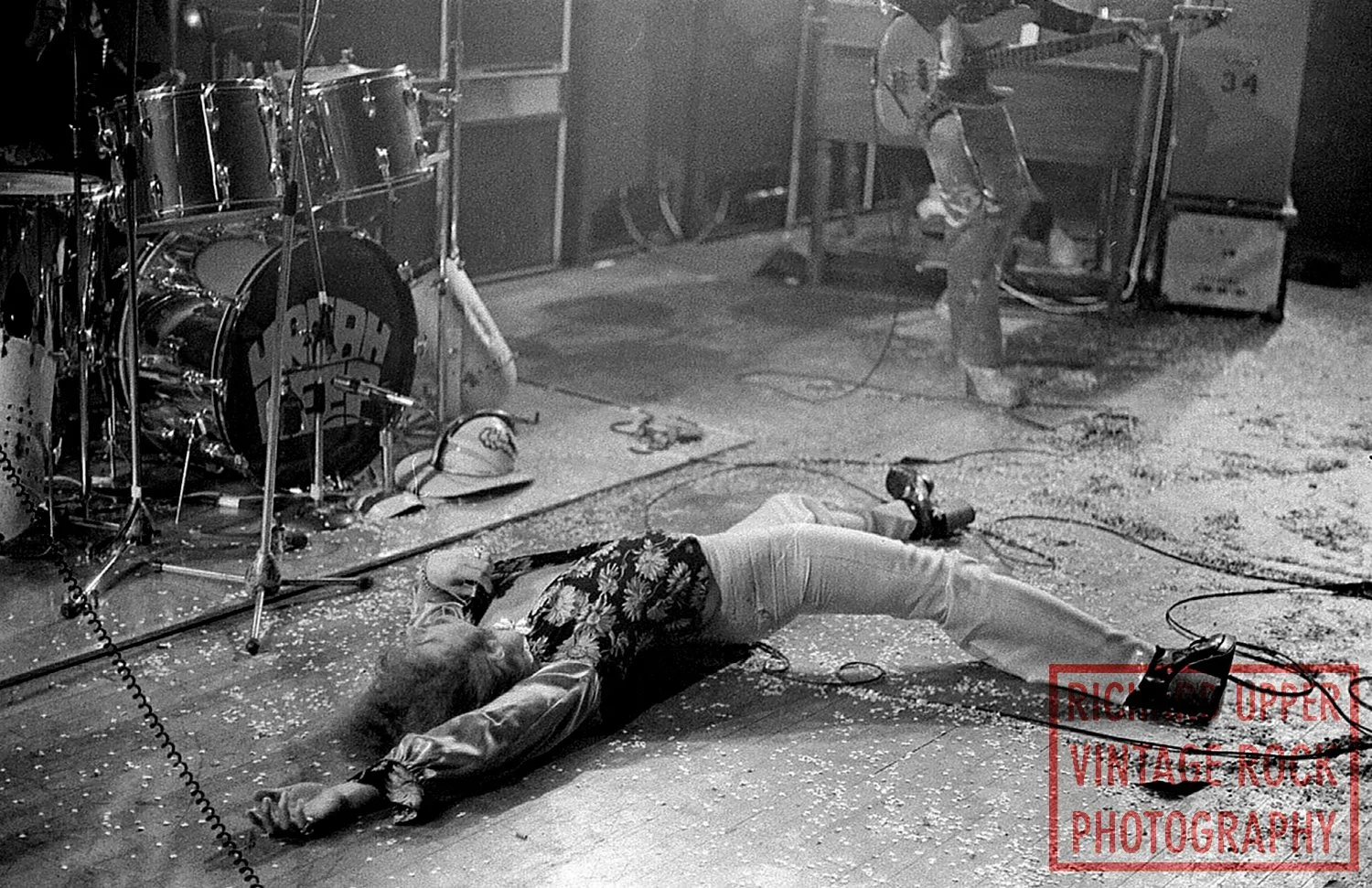 A person lying on the floor of a music studio or rehearsal space, surrounded by musical equipment including drums and an amplifier, with a person adjusting sound equipment in the background. The setting appears to be a rock or vintage music environme
