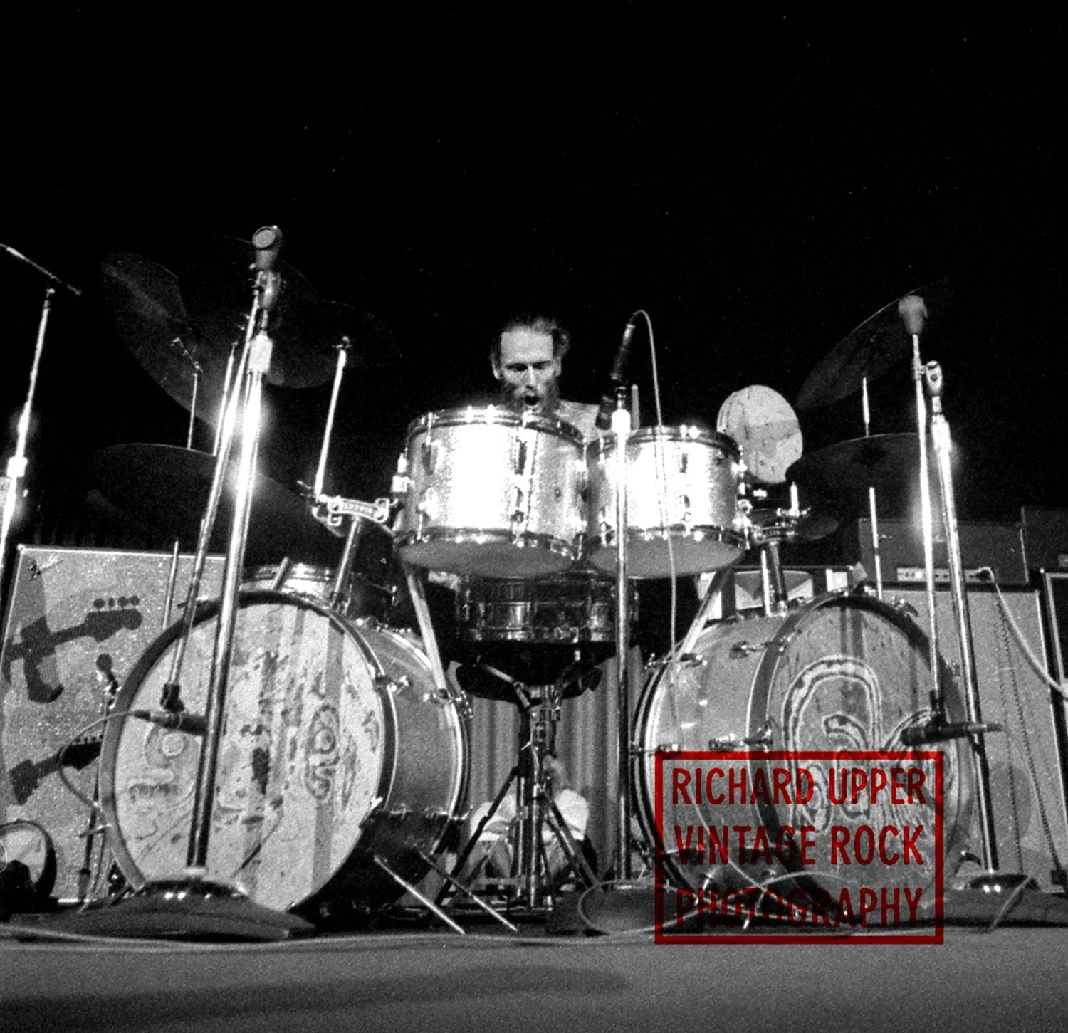 Black and white photo of a drummer behind a drum set on stage, with a sign reading 'Richard Upper Vintage Rock Photography' in front of the drums.
