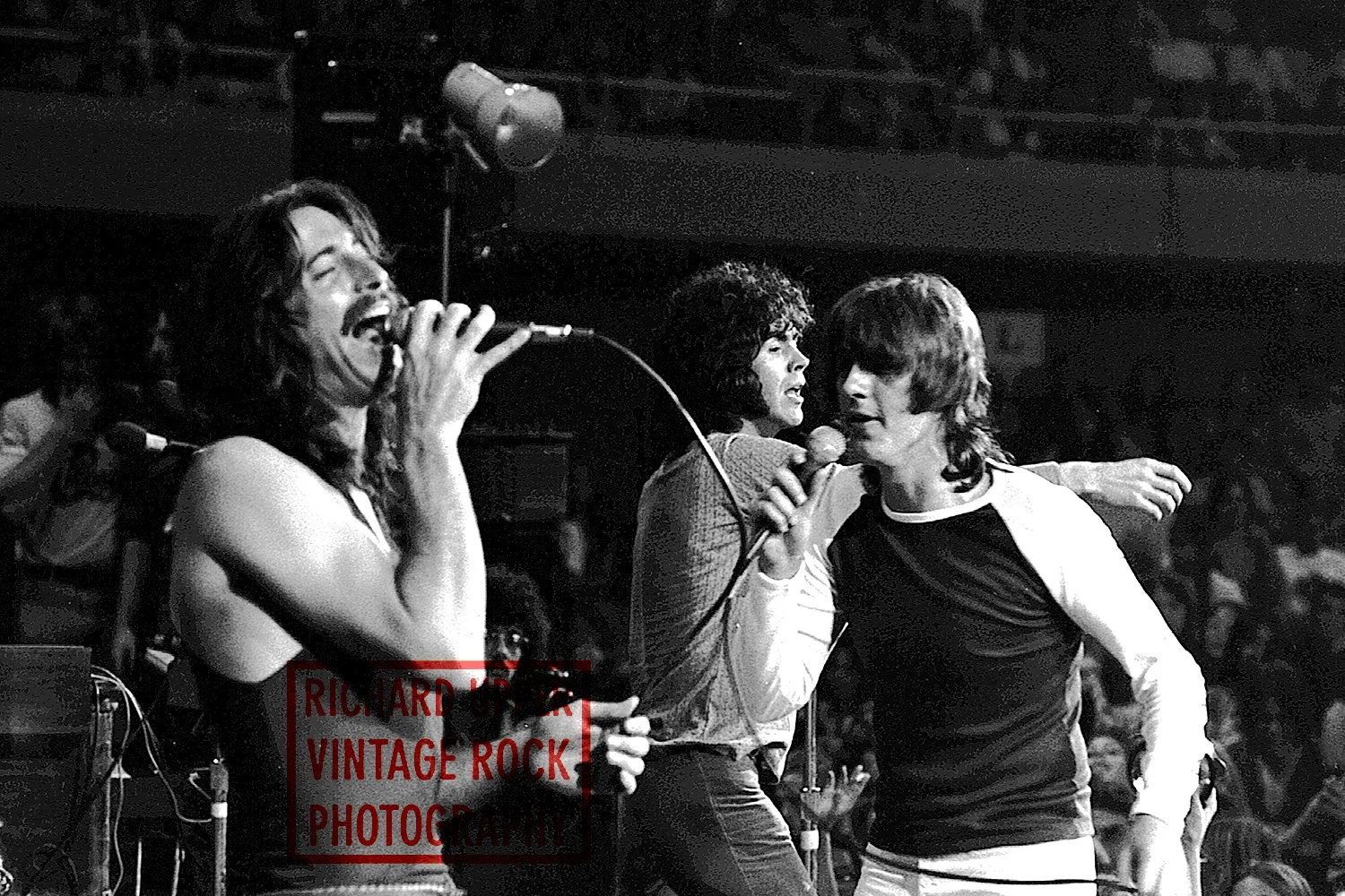 Black and white photo of three male singers performing on stage with microphones, singing passionately, with an audience in the background.