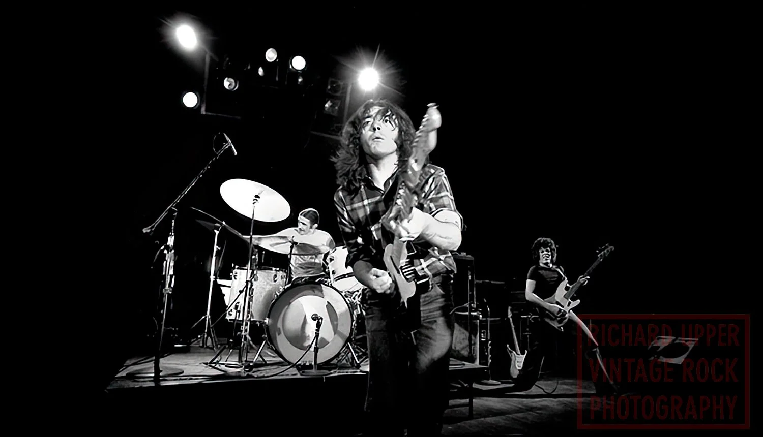 Black and white photo of a vintage rock band performing on stage, with a female bassist, a drummer, and a female guitarist. The bassist is in the foreground with a focused expression, holding a bass guitar, while the drummer and guitarist are in the 
