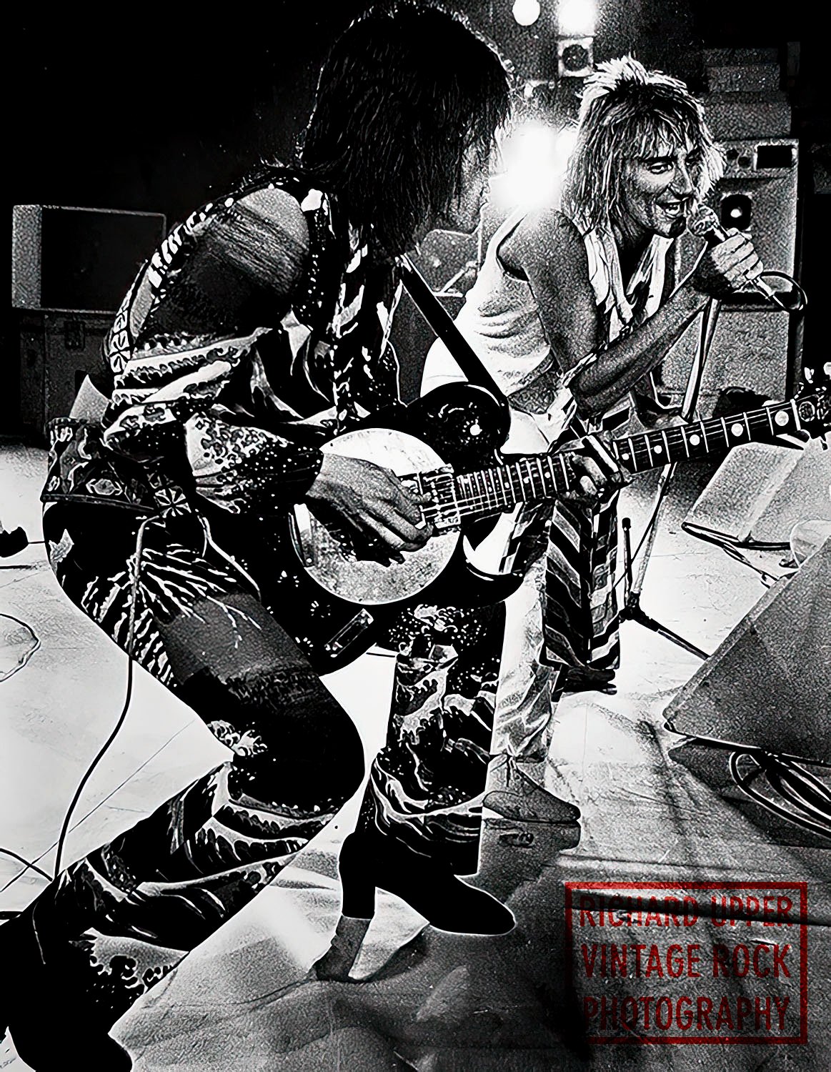 Two musicians performing with one playing guitar and the other singing into a microphone. The scene appears to be in a live music setting with equipment visible in the background.