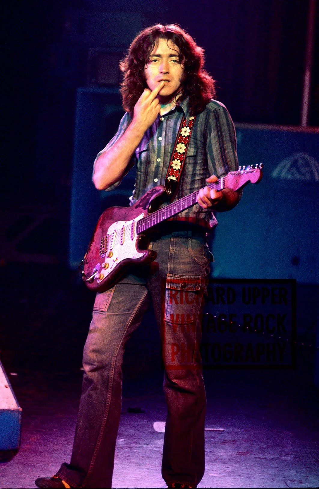 A young man with long curly hair playing an electric guitar on stage. He is wearing a plaid shirt and cargo pants, with a guitar strap decorated with daisies.