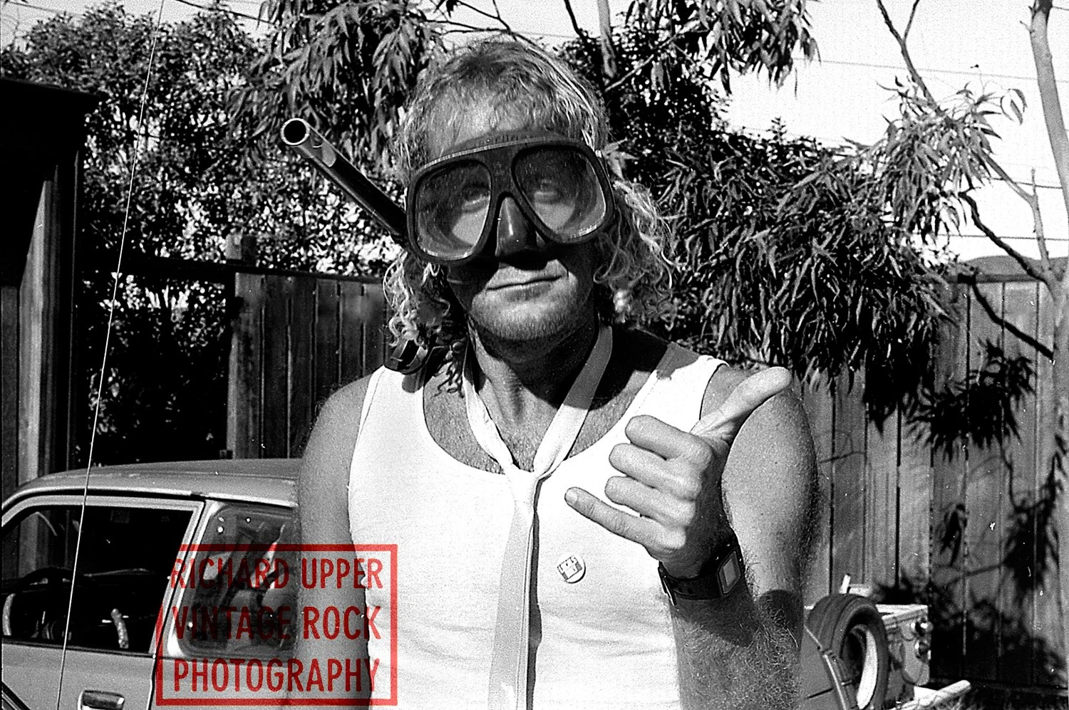 A man with curly hair wearing snorkeling gear, a sleeveless shirt, and a watch, making a rock and roll hand gesture with his right hand, outdoors with trees and a fence in the background.
