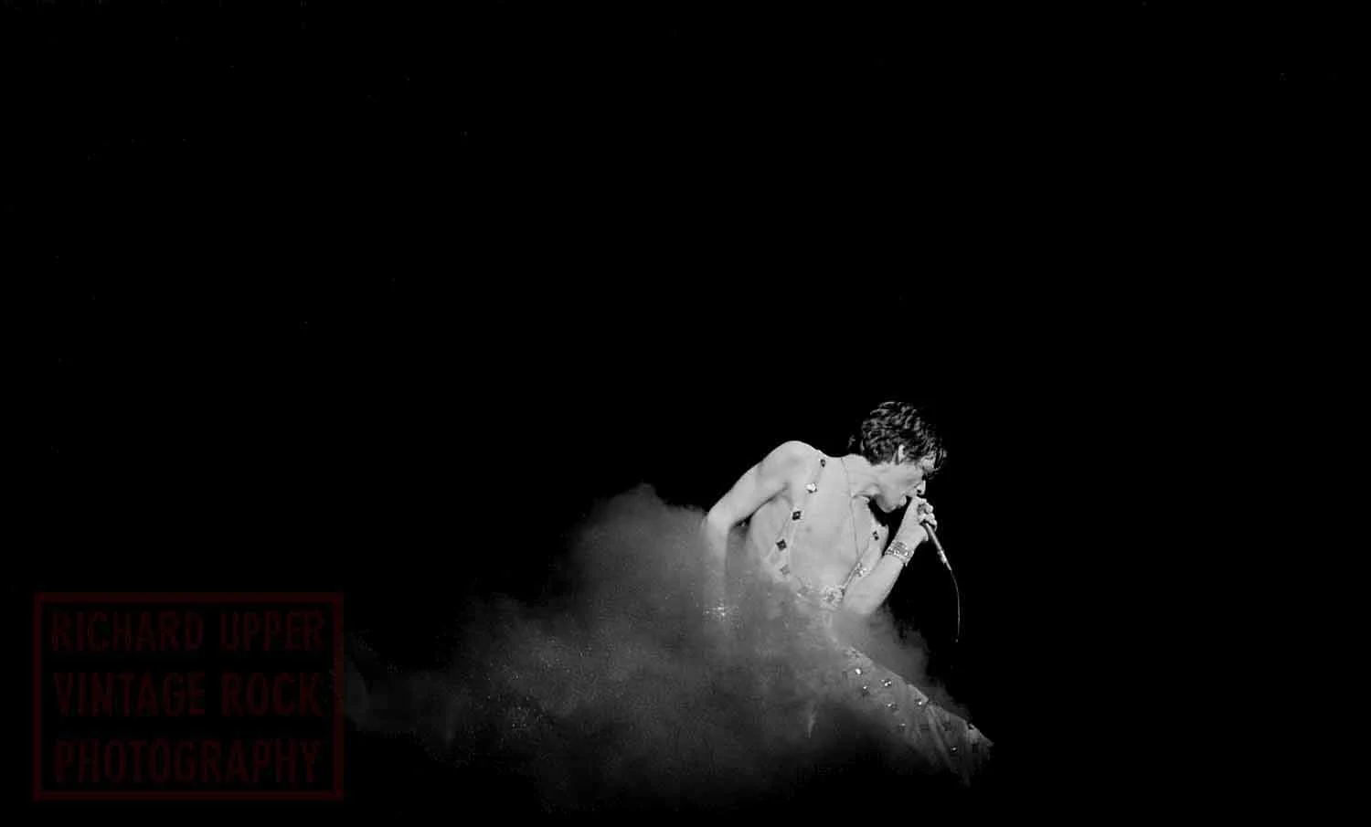 Black and white photo of a singer performing on stage, standing with a microphone, with a dark background.