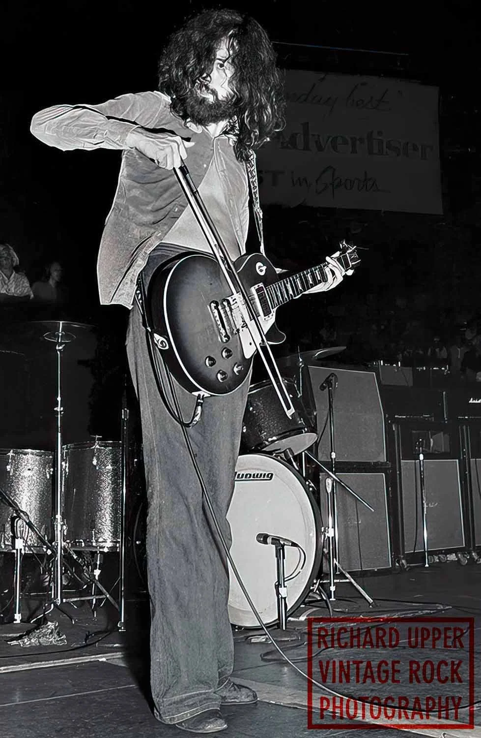 A musician with long hair and beard playing an electric guitar on stage, with drums and amplifiers in the background, in a black-and-white photo.