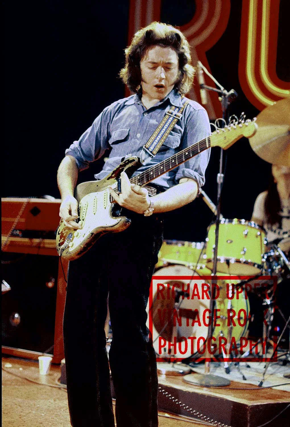 A person playing an electric guitar on stage, wearing a blue button-up shirt with rolled-up sleeves, with a drum set and microphone in the background. The stage has a retro style with neon lights.