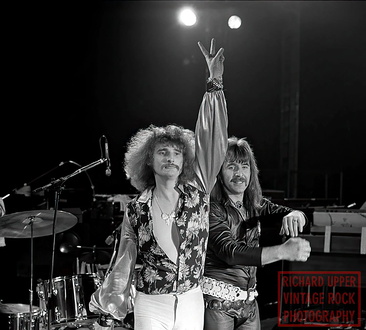 Black and white photo of two men, one with curly hair and a mustache raising his hand, the other with straight hair and a mustache, both posing on a stage with musical instruments in the background. The bottom right corner has a red box with the text