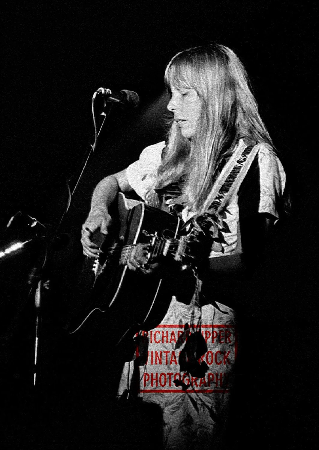 A woman with long hair playing an acoustic guitar on stage, with a microphone in front of her, in black and white.