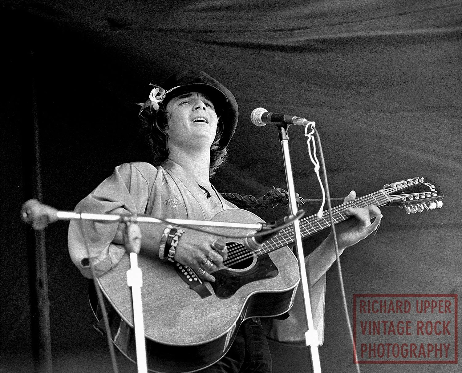 A woman wearing a hat and vintage-style clothing plays an acoustic guitar and sings into a microphone backstage, with a dark backdrop.