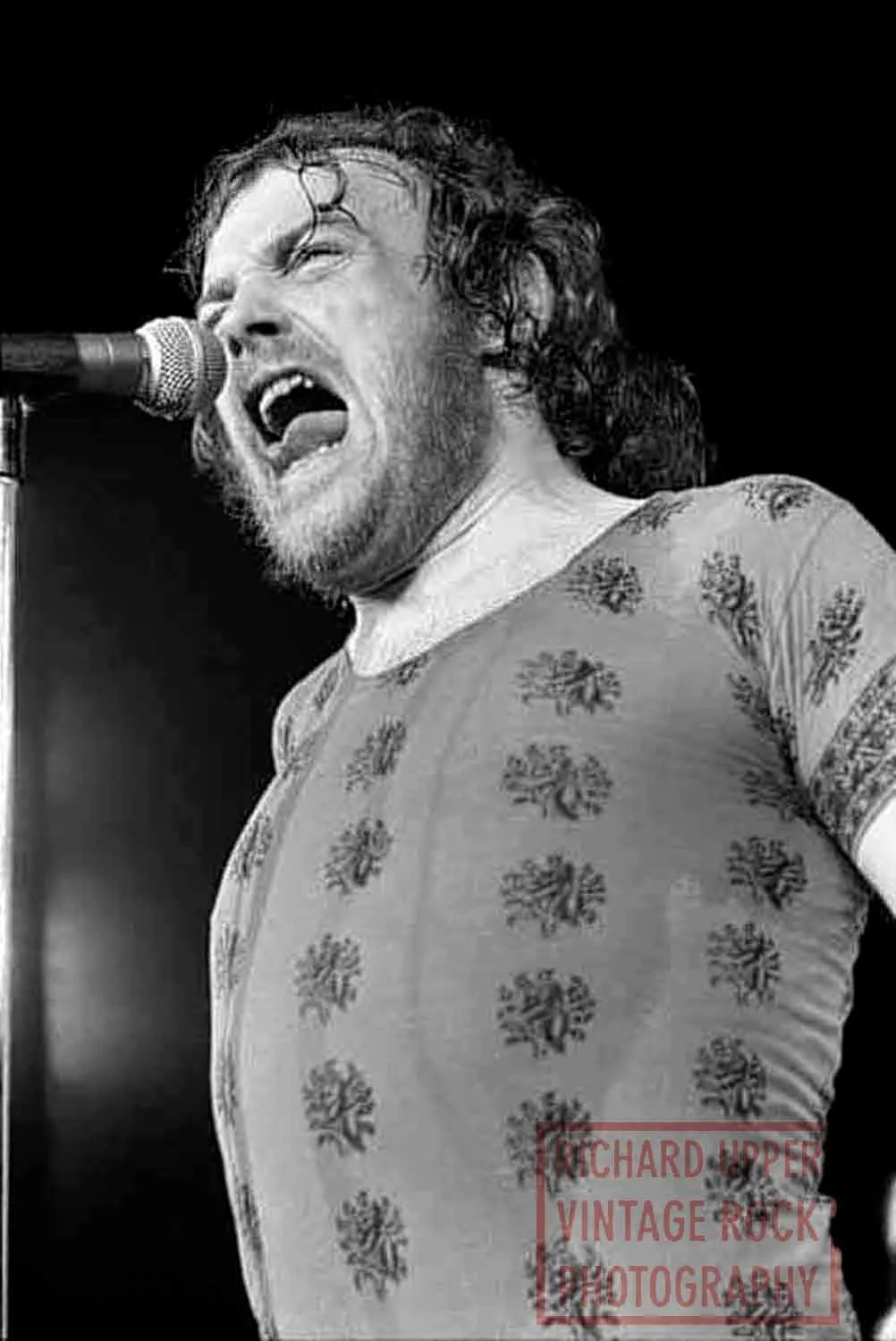 Black and white photo of a man singing passionately into a microphone on stage, wearing a patterned t-shirt.