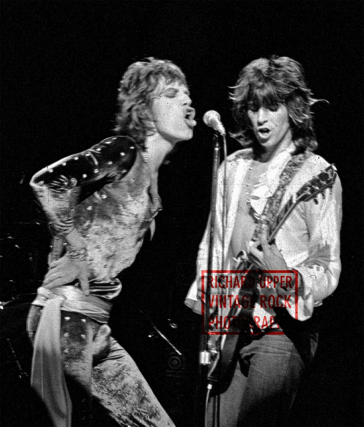 Black and white photo of two male musicians singing into a microphone. The musician on the left is wearing a patterned shirt with rolled-up sleeves and patterned pants, with curly hair. The musician on the right has long wavy hair, wearing a light-co