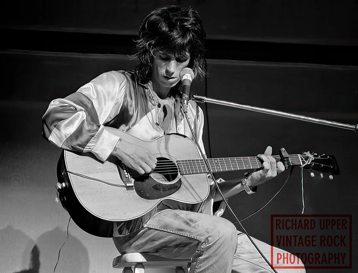A person with shoulder-length hair playing an acoustic guitar and singing into a microphone on stage, in black and white.