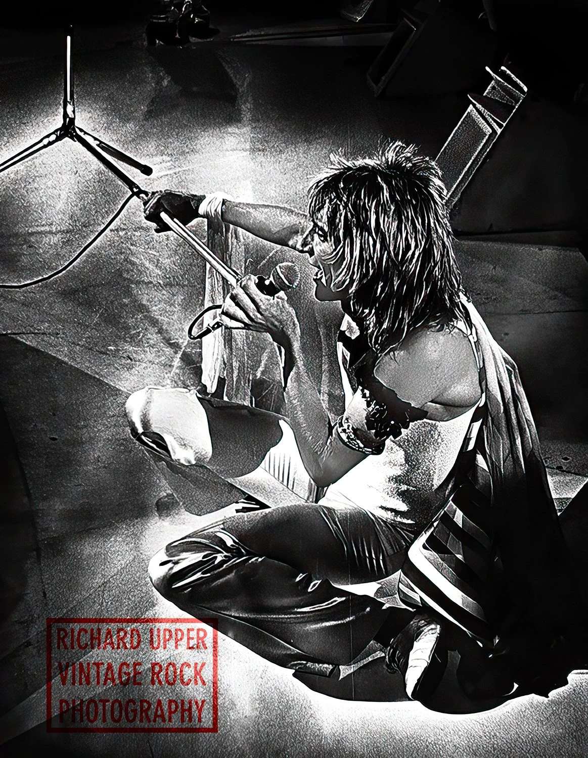 A black and white photograph of a person singing into a microphone, sitting on a chair with a seatbelt, in a studio setting. The person has wild, shoulder-length hair, is wearing a sleeveless top, and is surrounded by professional studio equipment. T
