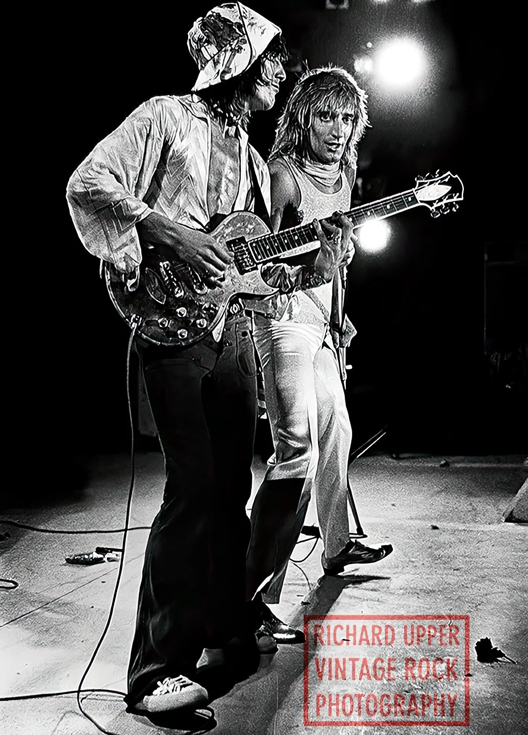 Two musicians on stage, one playing an electric guitar and the other singing, with bright stage lights in the background, black and white photo.