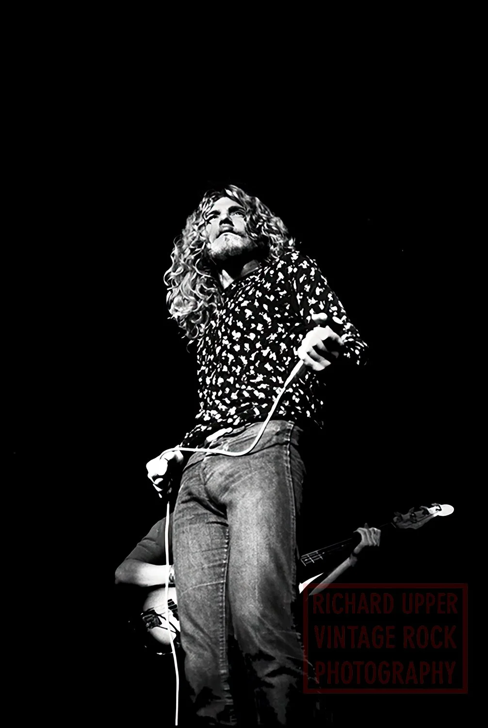 A black and white photo of a male singer with long, curly hair, wearing a patterned shirt, holding a microphone, and performing on stage. A guitarist is partially visible in the background.