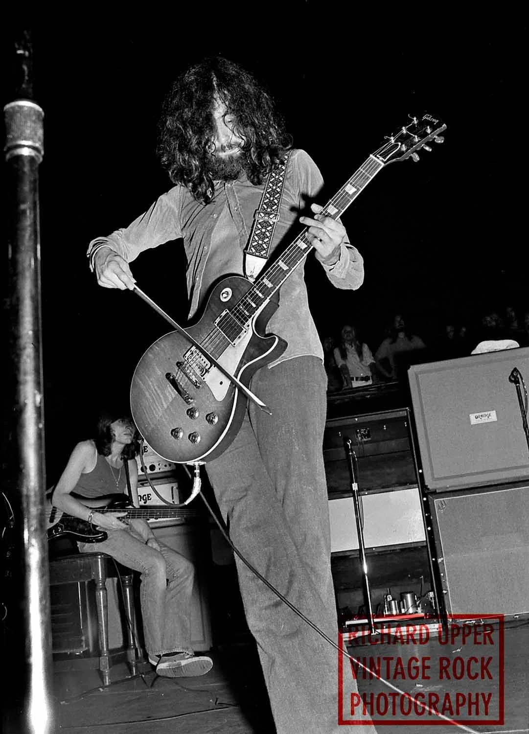 Musician with curly hair playing electric guitar on stage, with a bassist seated in the background, black and white photo.