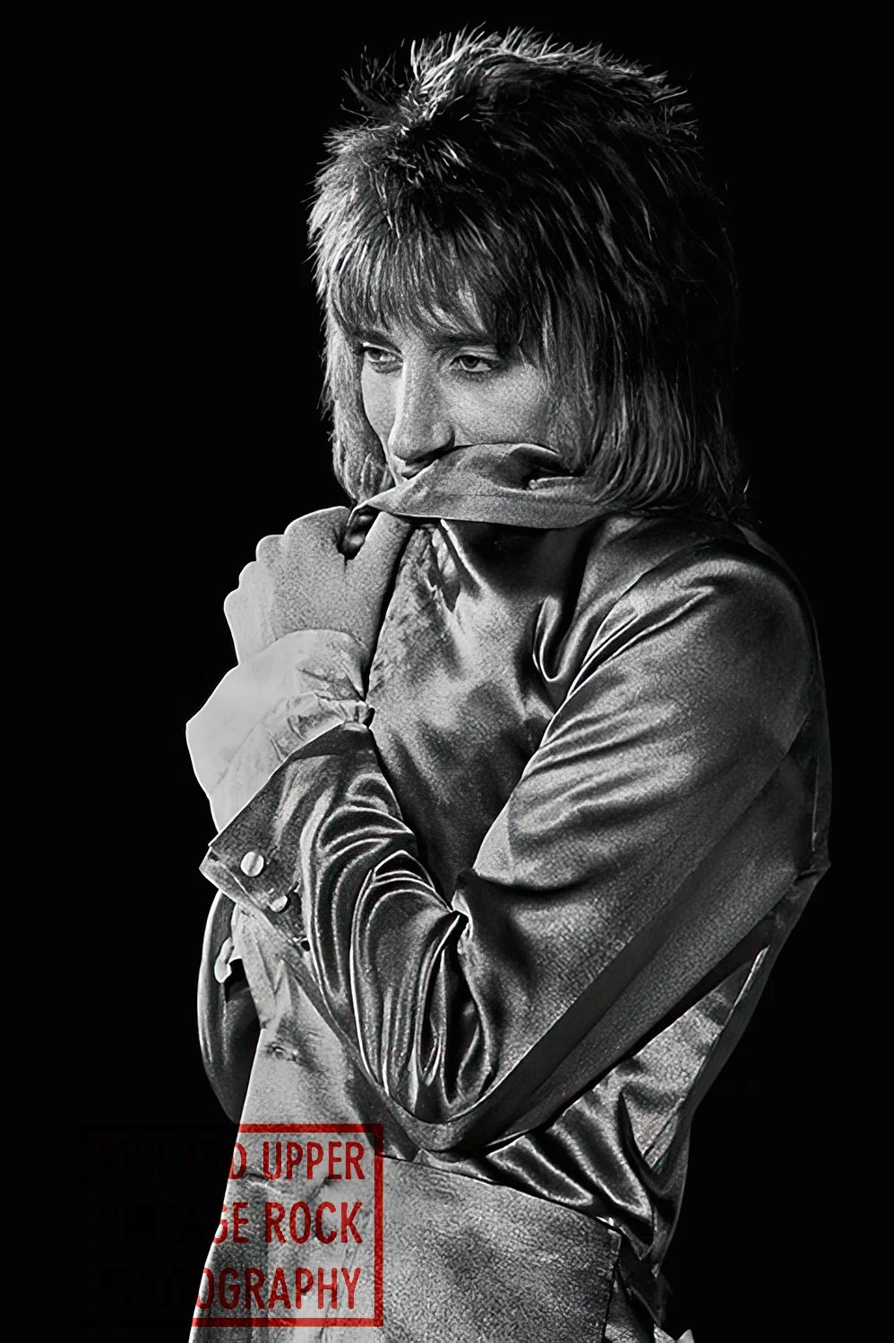 Black and white portrait of a man with long hair, wearing a leather jacket, with his hand near his face, looking contemplative.