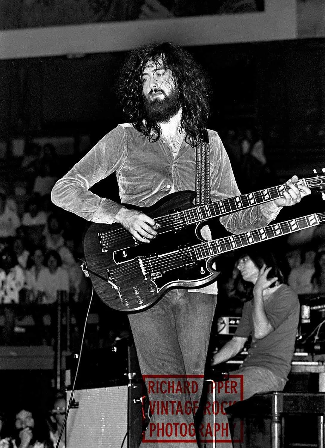 A musician with curly hair and a beard playing a double-neck guitar on stage at a concert, with an audience in the background.