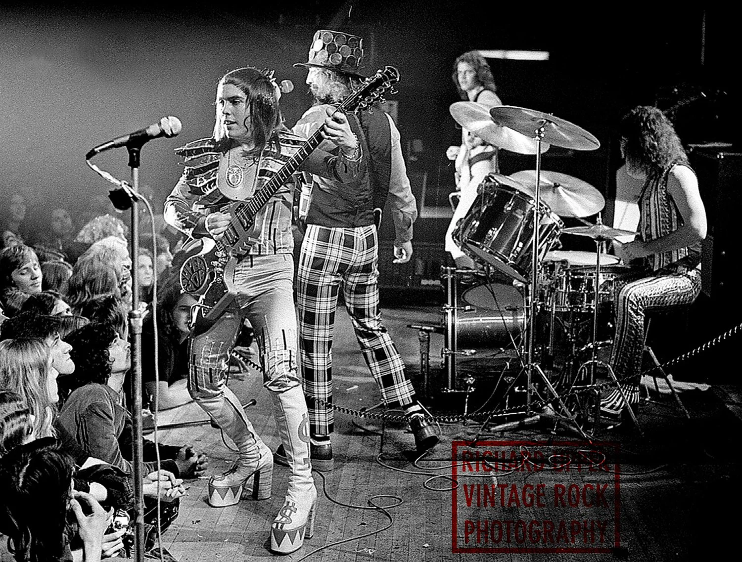 Black and white photo of a vintage rock concert with female lead singer holding a guitar, a female drummer, and audience members watching attentively.