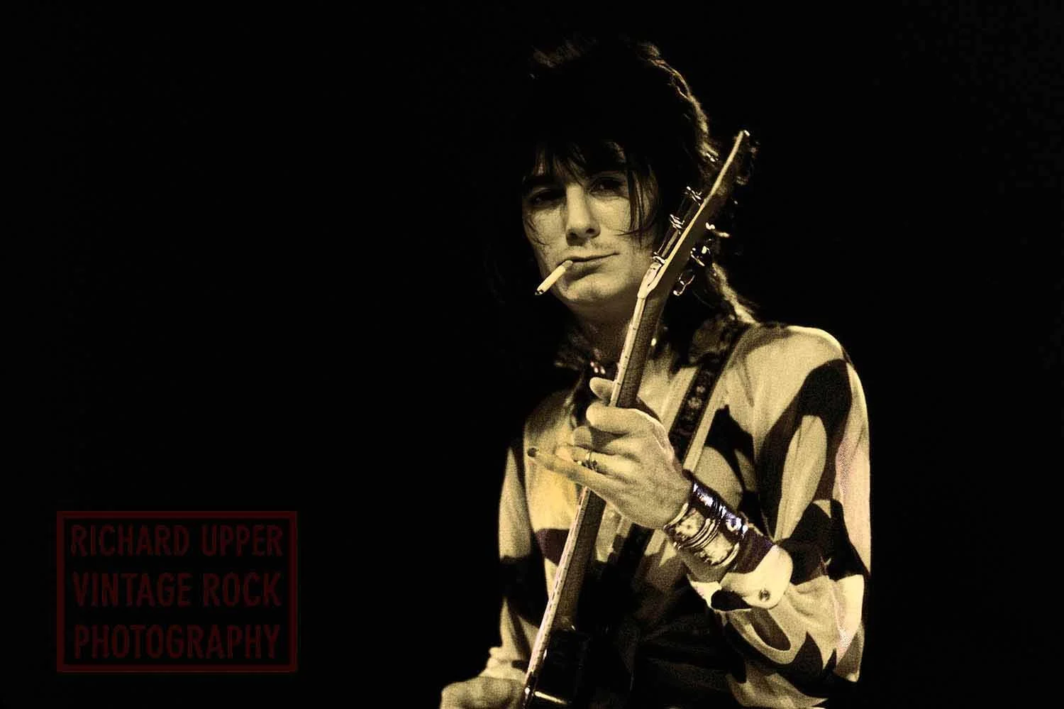 Black and white photo of a man with dark hair playing an electric guitar, with a cigarette in his mouth, wearing a patterned shirt, and wearing multiple bracelets on his wrist. The photo has a logo that reads "Richard Upper Vintage Rock Photography" 