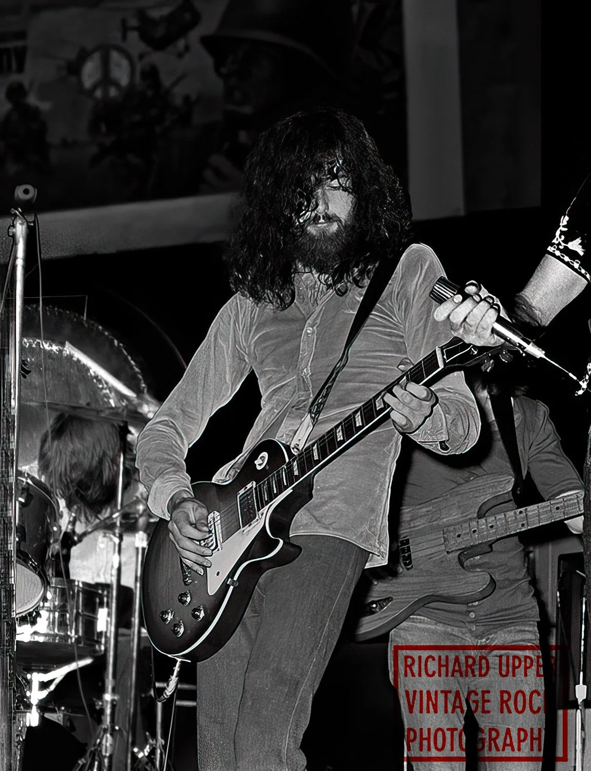Black and white photo of a guitarist with long curly hair and beard playing an electric guitar on stage, with a drummer partially visible in the background.