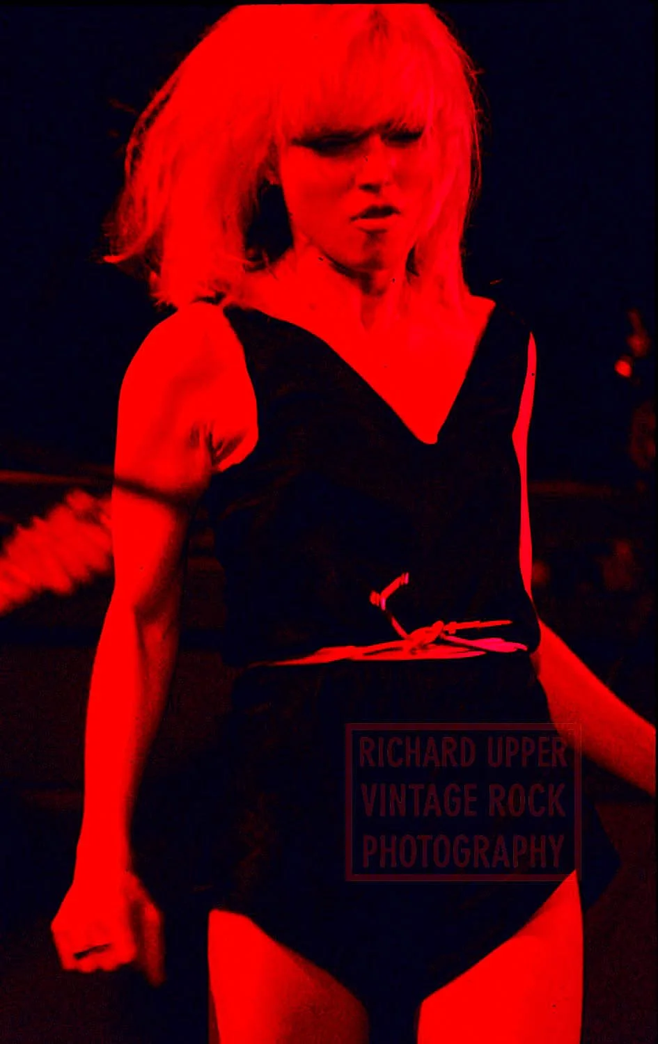 A woman with tousled blonde hair, dressed in a black sleeveless dress tied at the waist, posing against a dark background, bathed in red lighting.