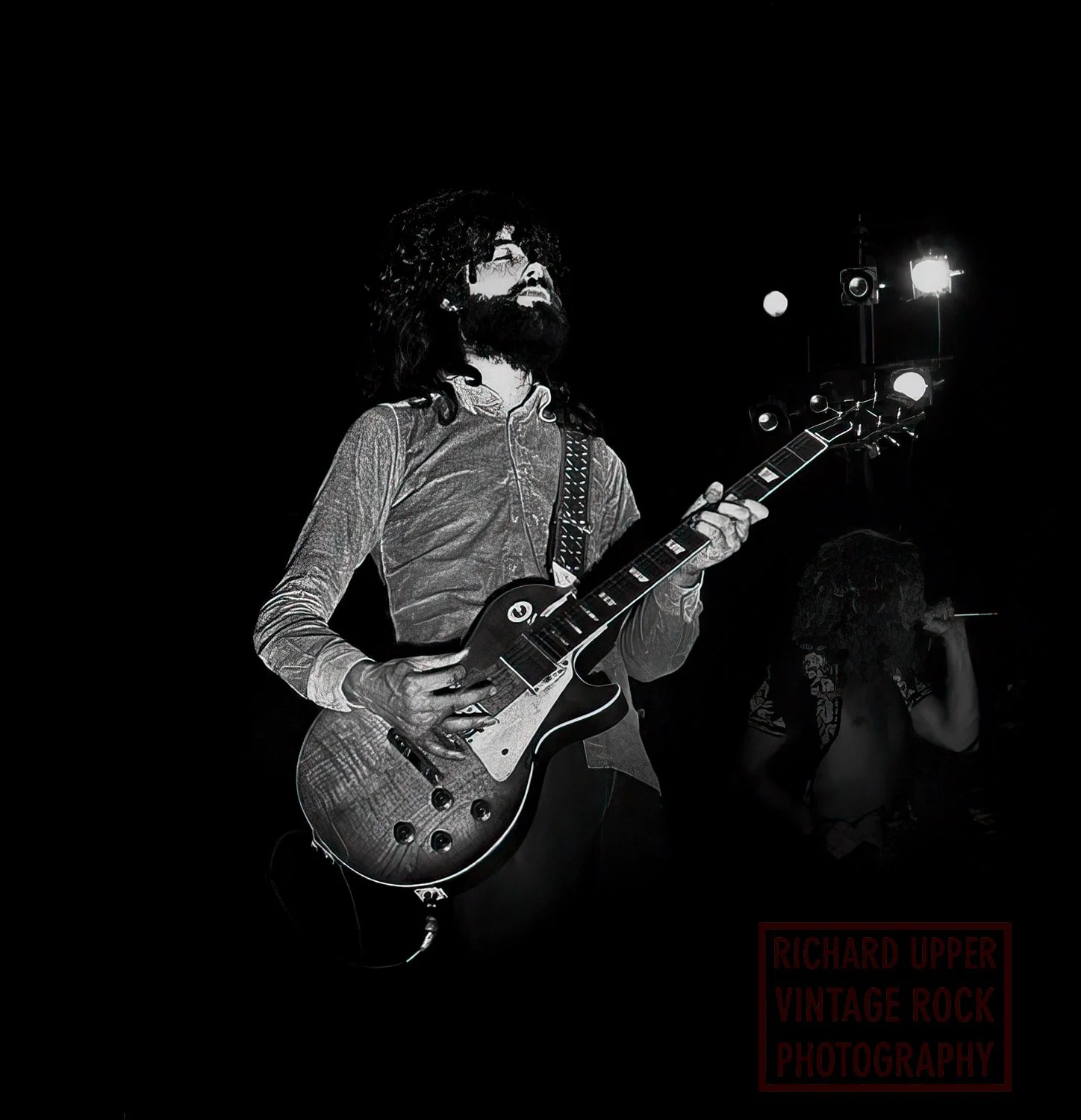 A man with long hair and a beard playing an electric guitar on stage, with stage lights in the background. Another person is in the background, possibly singing or speaking into a microphone.