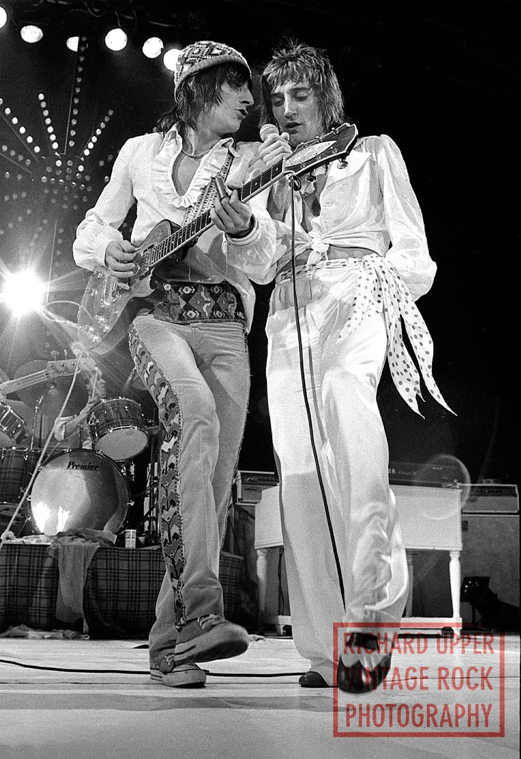 Two musicians performing on stage with a guitar and microphone, dressed in vintage clothing, with drums and stage lights in the background.
