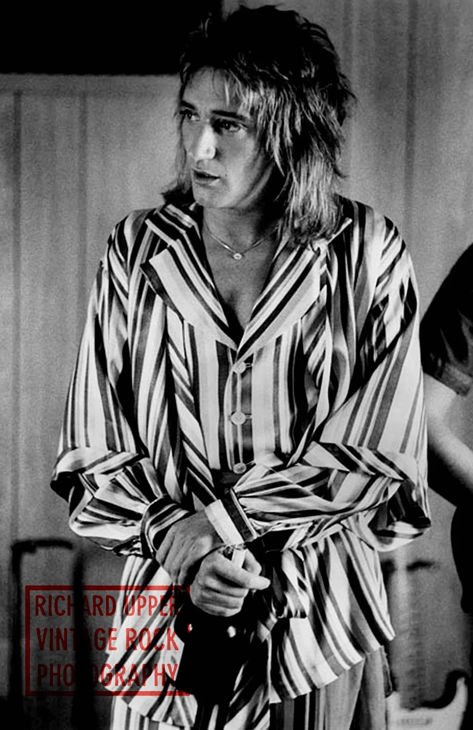 Black and white photo of a woman with shoulder-length, layered hair wearing striped pajamas, holding a microphone, with a partial name overlay reading 'RICHARD UPPER VINTAGE ROCK PHOTOGRAPHY'.