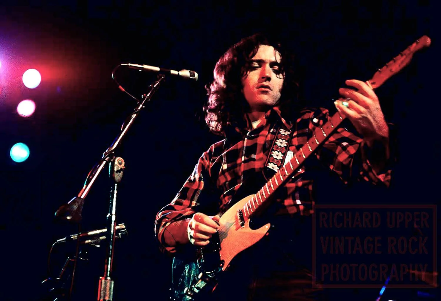A musician with long curly hair playing an electric guitar on stage, wearing a red and black plaid shirt, with microphone set up in front of him and colorful stage lights in the background.