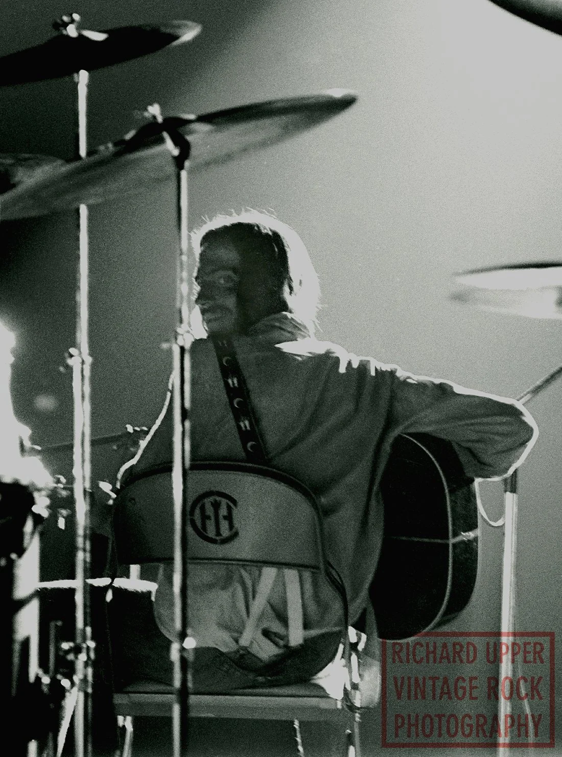 A black and white photo of a man with long hair playing an acoustic guitar next to a drum set.