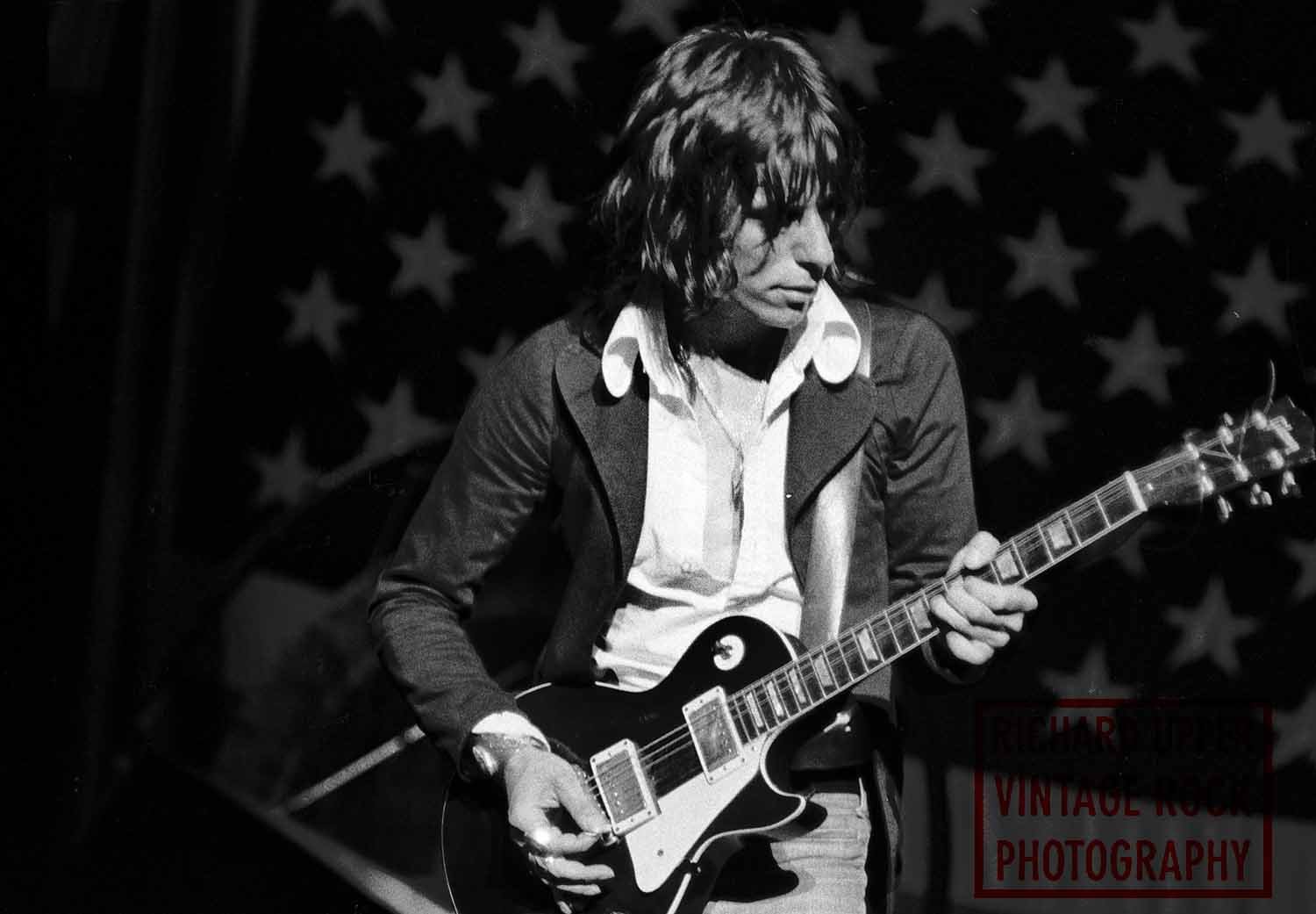 Black and white photo of a man playing an electric guitar on stage with an American flag backdrop.