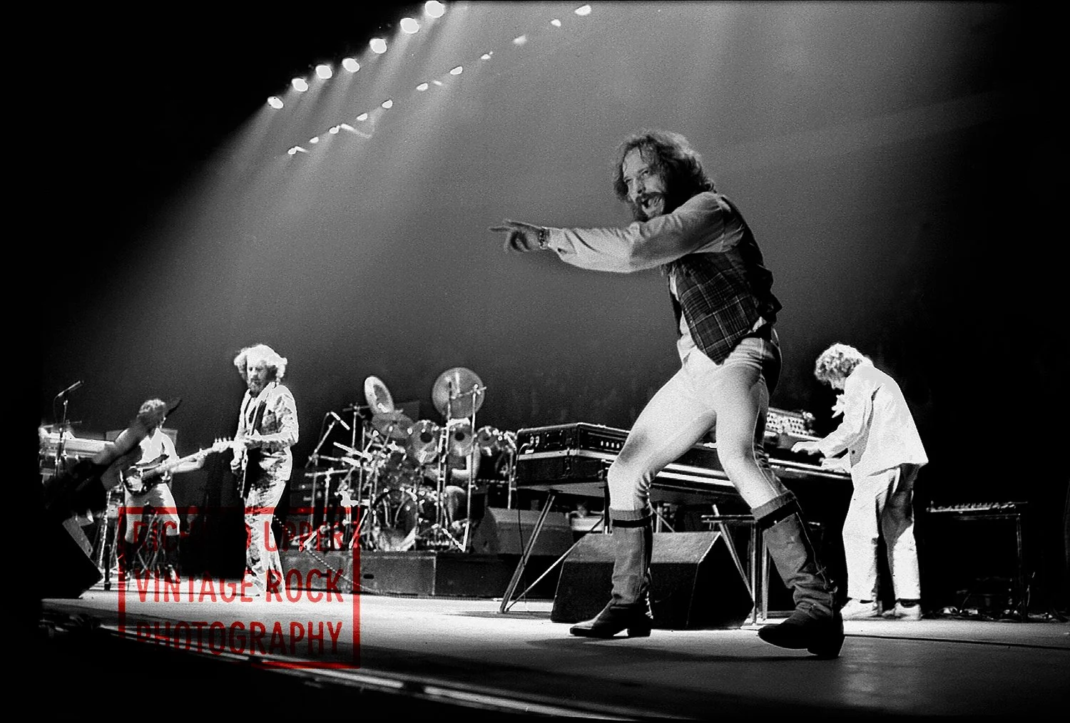 A black and white photo of a vintage rock band performing on stage with a lead vocalist in the foreground pointing towards the audience, surrounded by guitars, drums, and keyboards.