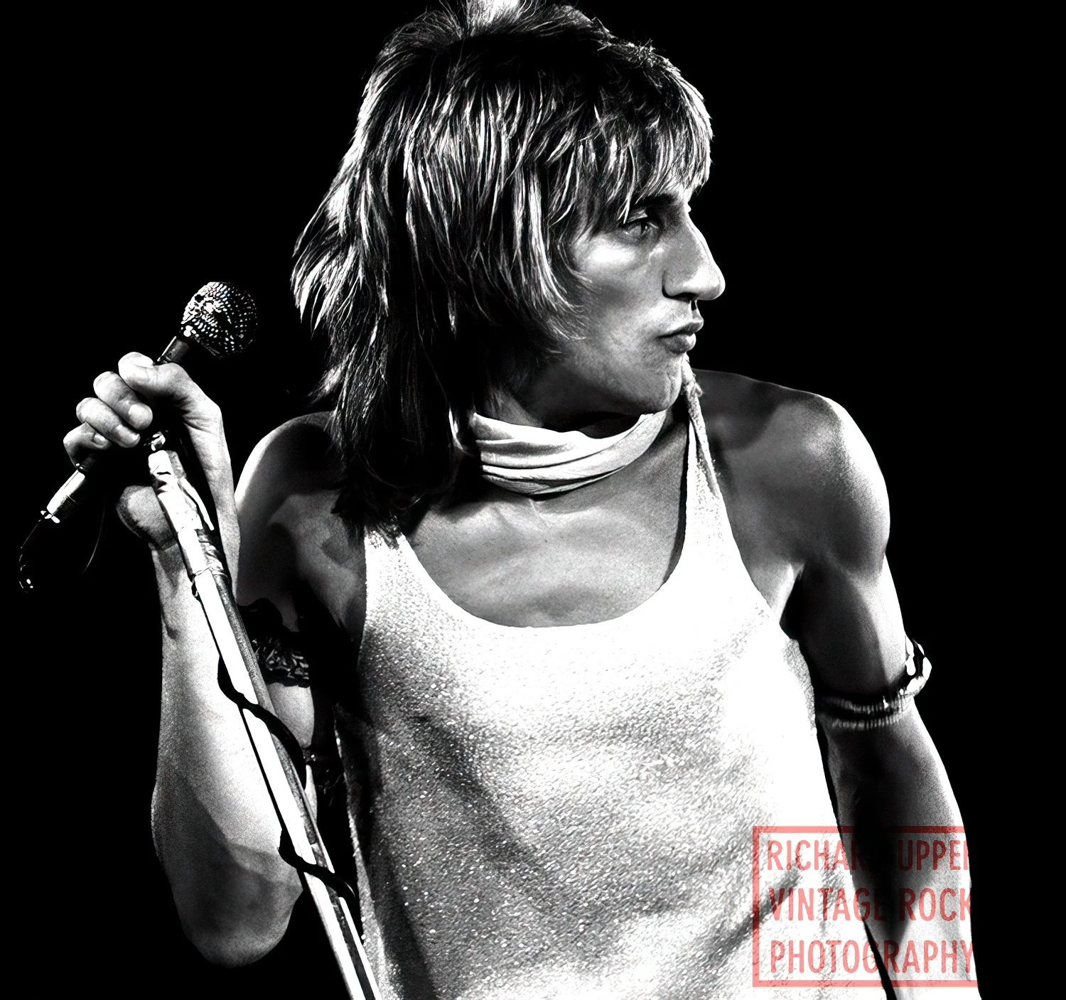 Black and white photo of a woman singing into a microphone, wearing a sleeveless top, with shoulder-length hair, and a scarf around her neck, against a dark background.