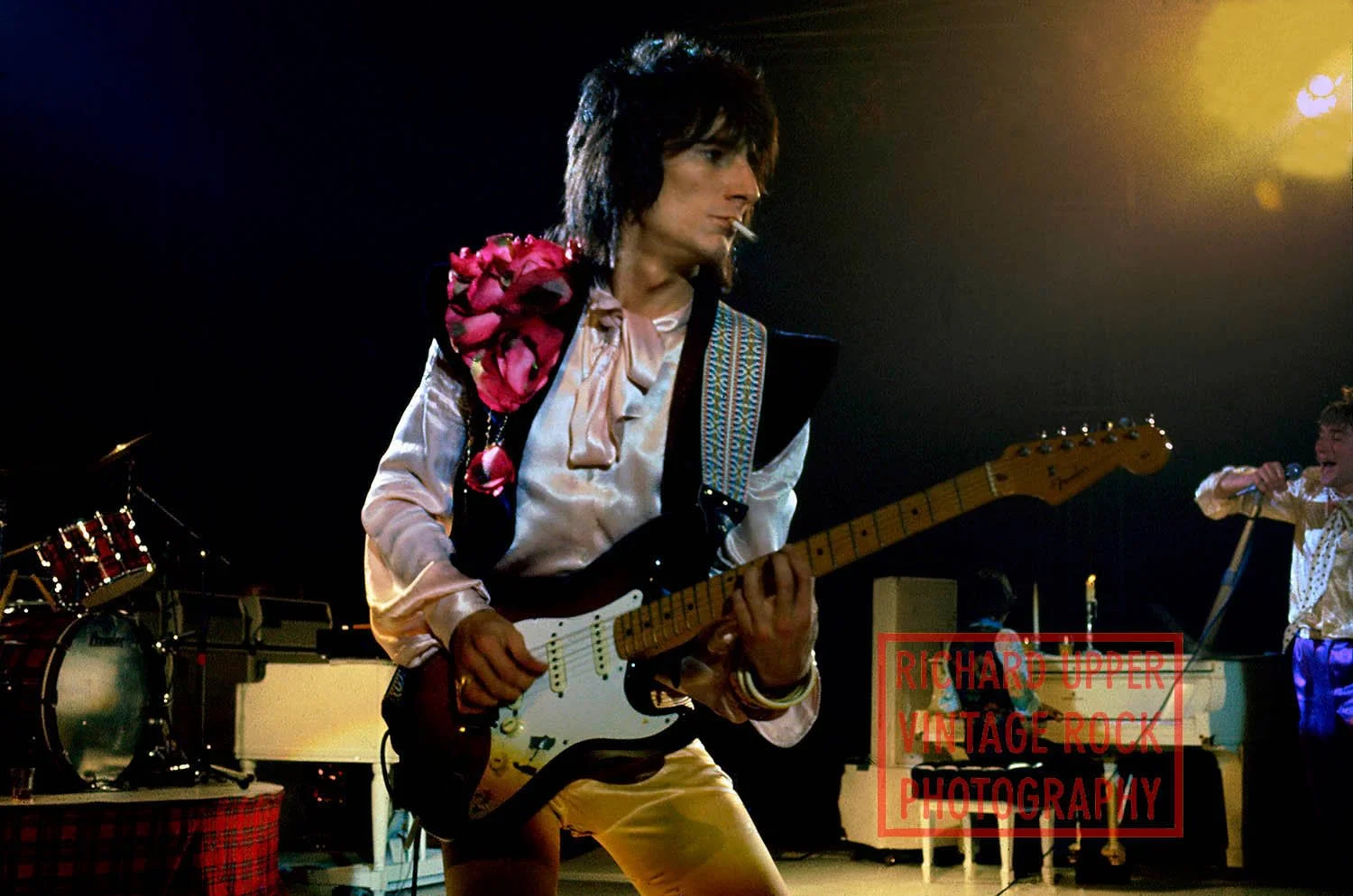A musician playing an electric guitar on stage, wearing vintage rock attire with a flower on his shoulder, and a band member singing into a microphone in the background.