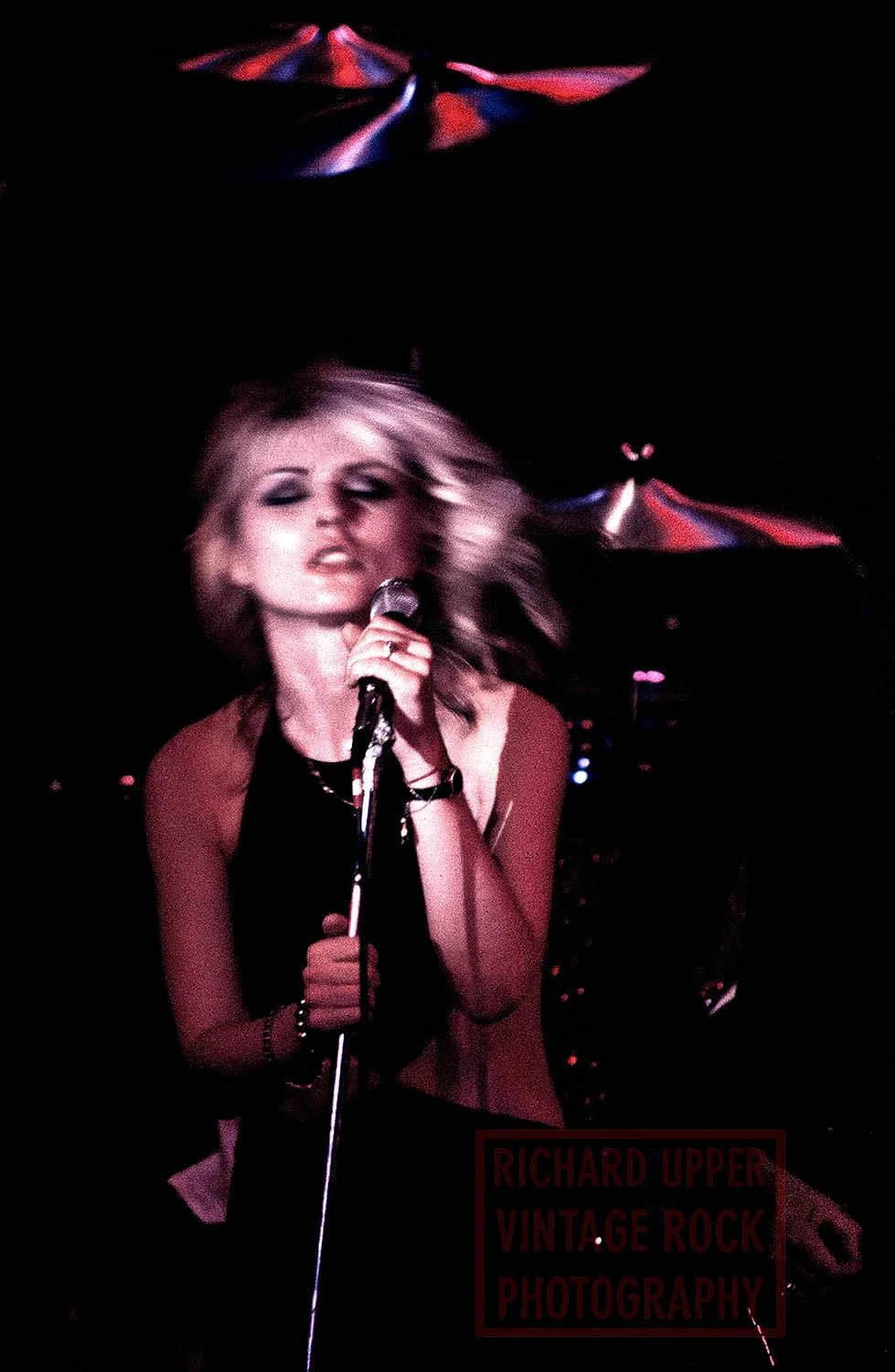 A woman singing into a microphone on stage with colorful stage lights and a dark background. The photo appears slightly blurry with motion, capturing her energetic performance.