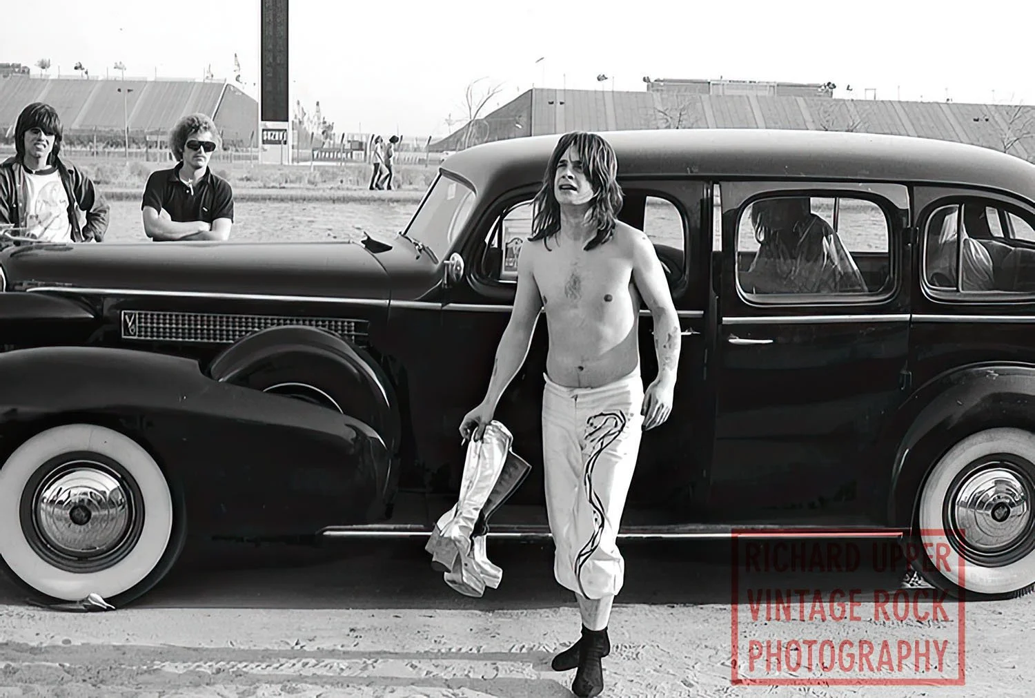 A shirtless man with long hair and tattoos walking away from a vintage black car, holding a shoe, with two people in the background and an outdoor setting.
