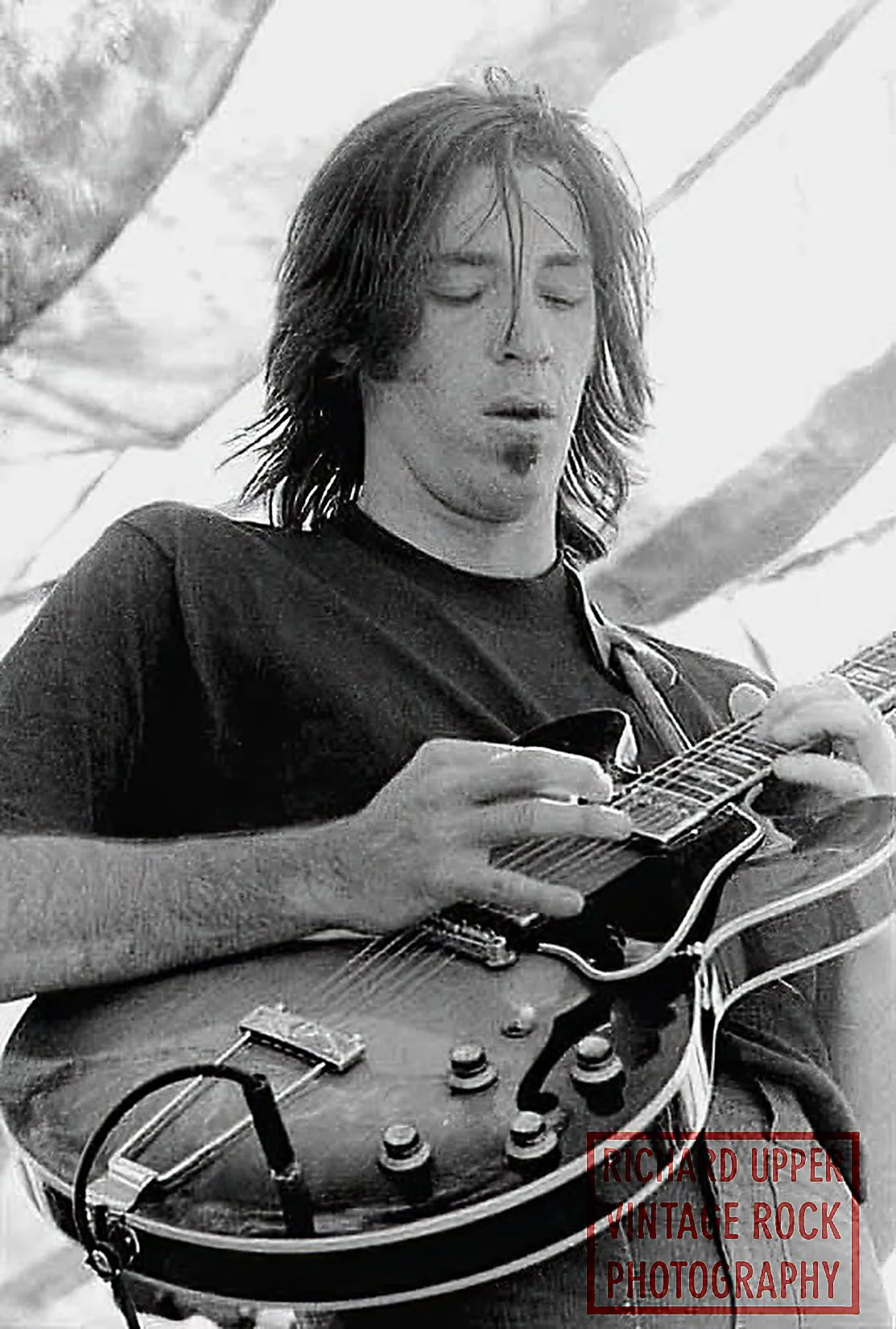 Black and white photo of a young man with shoulder-length hair playing an electric guitar, eyes closed, with a background that appears to be a tent or outdoor setting, and the text 'RICHARD UPPEN VINTAGE ROCK PHOTOGRAPHY' in the bottom right corner.