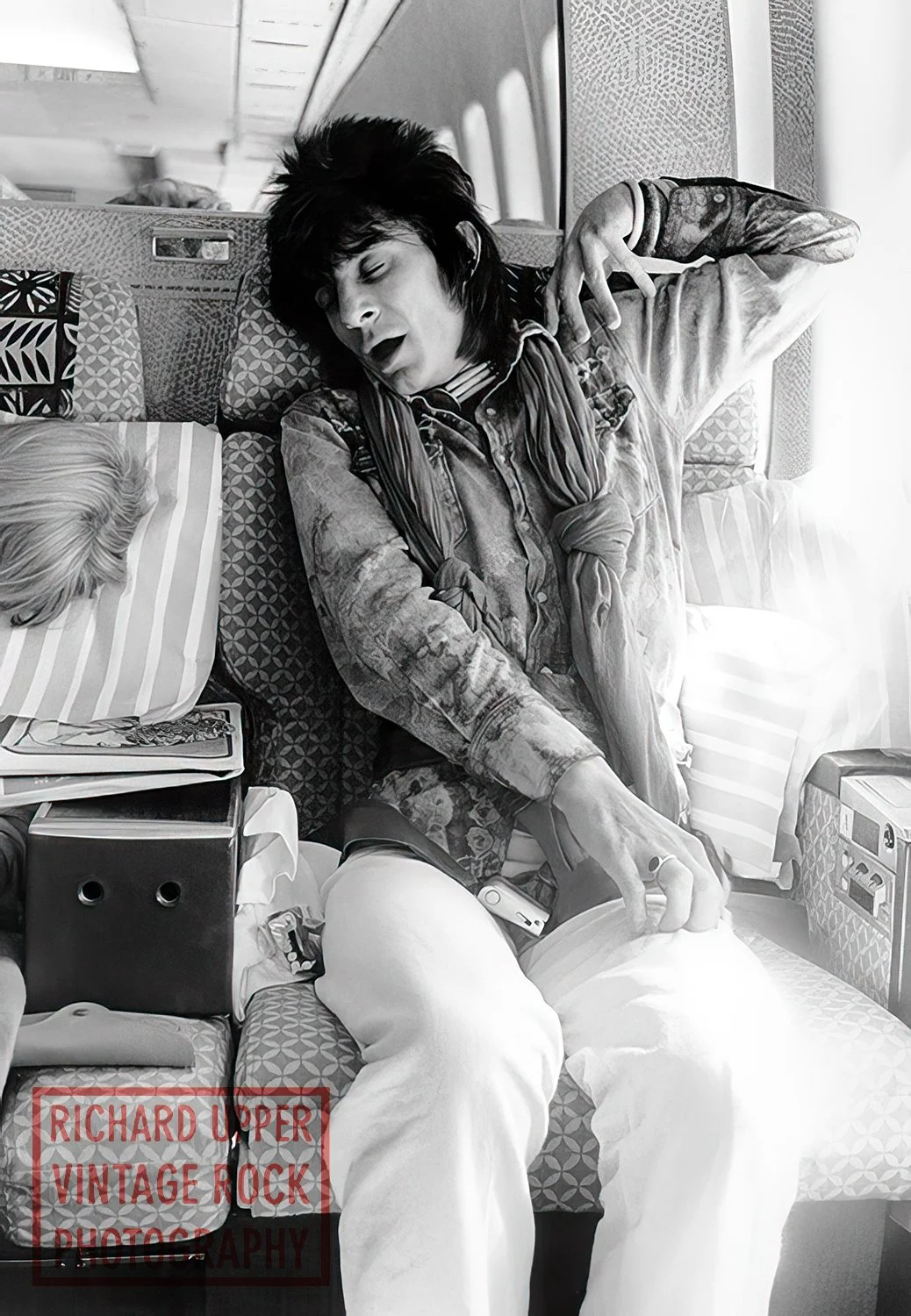 A woman with dark, voluminous hair, wearing a patterned shirt and light-colored pants, is sleeping on a bus seat with her head resting on a pillow. Her arm is extended overhead and her mouth is slightly open. An umbrella and other personal items are 