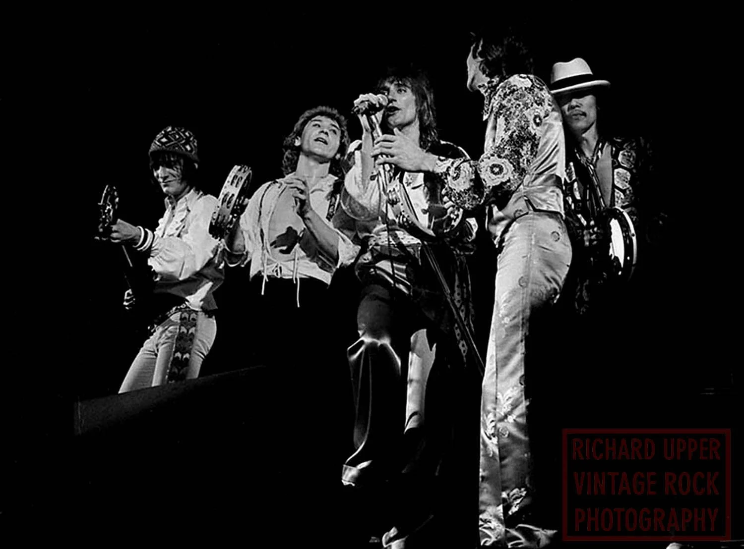 Black and white photo of five musicians performing on stage, with one playing guitar and others singing into microphones, dressed in vintage rock style clothing.