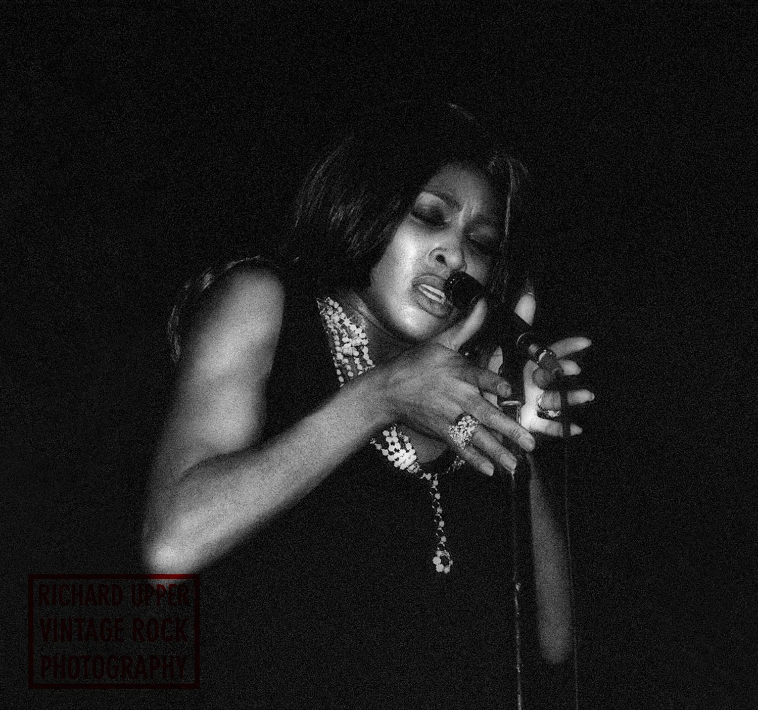 Black and white photo of a woman singing passionately into a microphone, wearing jewelry and a sleeveless dress.