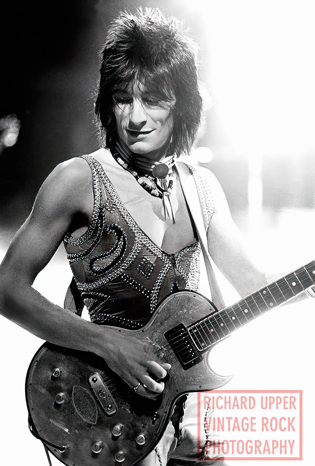 Black and white photo of David Bowie performing with an electric guitar, wearing a sleeveless, embellished top, with short, spiky hair, on stage under bright lights.